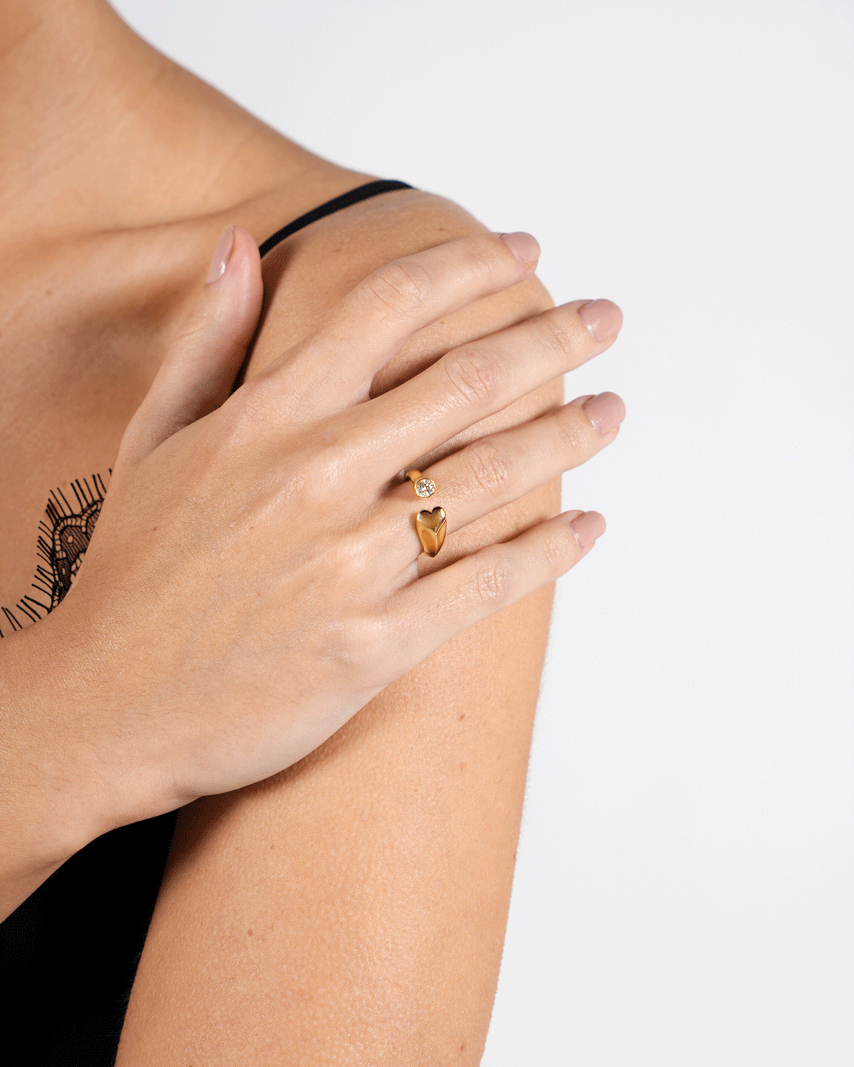 Close-up of a woman's hand resting on her shoulder, displaying a unique gold ring with a heart motif and a round diamond on her ring finger. She wears a black sleeveless top, emphasizing the elegance of the ring against her skin.