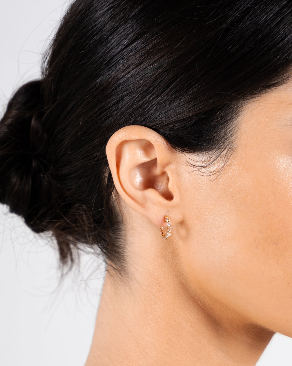Close-up view of a woman's ear with dark hair styled in a neat bun, wearing elegant marquise-cut diamond huggie earrings. The earrings feature a vintage design with a golden glow, accentuating her sophisticated and polished look.